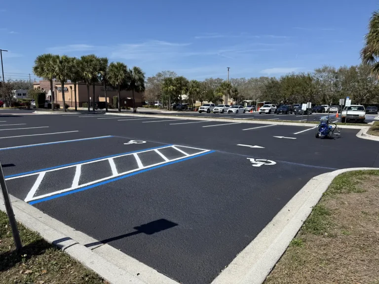 Freshly sealcoated commercial parking lot with crisp white striping and blue ADA handicap markings at a Tampa Bay plaza. Wide view showing jet-black sealcoat finish across the full lot. Professional parking lot sealcoating by Radiant Striping, veteran-owned with 212 five-star Google reviews. Serving Tampa, St. Petersburg, Clearwater, and all of Tampa Bay. (813) 448-1252.